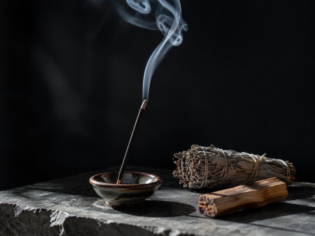 Incense smoke curling above a ceramic holder beside sage and palo santo on stone